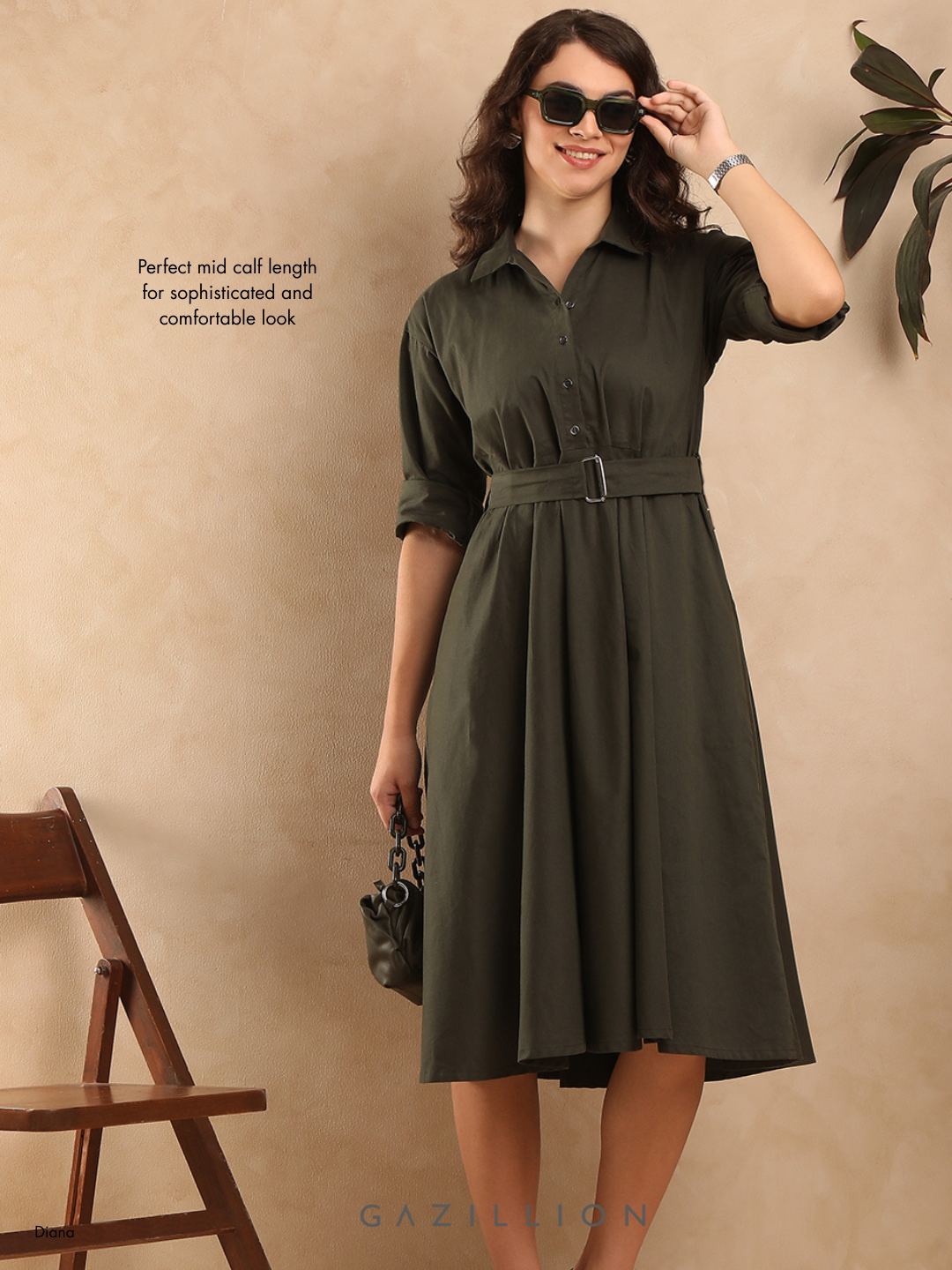 Woman wearing a green dress with a belt, standing against a beige wall with a wooden chair and plant.
