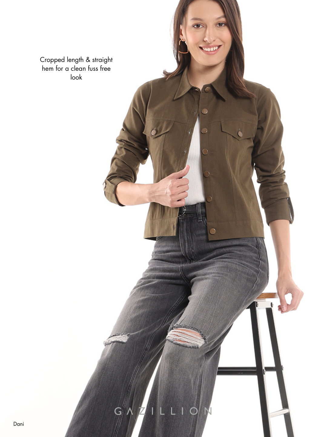 Woman wearing a brown jacket and gray jeans sitting on a stool with a white background